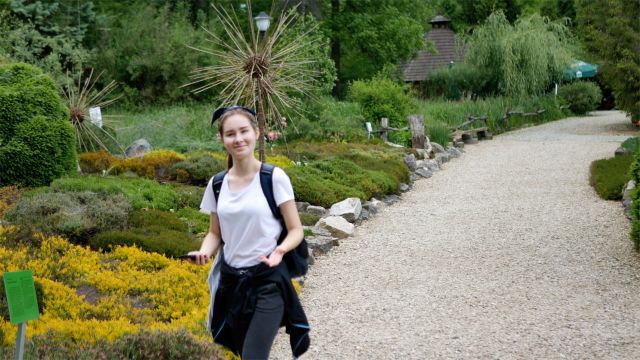 Uczestniczka wyjazdu w arboretum Bolestraszyce. Uśmiechnięta dziewczyna w białej bluzce, z czarnym plecakiem idzie ogrodową alejką.