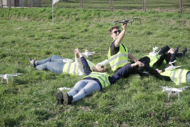 Kursanci z dronami. Jeden, pośrodku siedzi na trawie, reszta leży z rękami wyciągniętymi do góry.