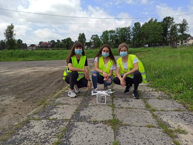 Trzy uczestniczki kursu w żółtych kamizelkach odblaskowych, kucające przy dronie.