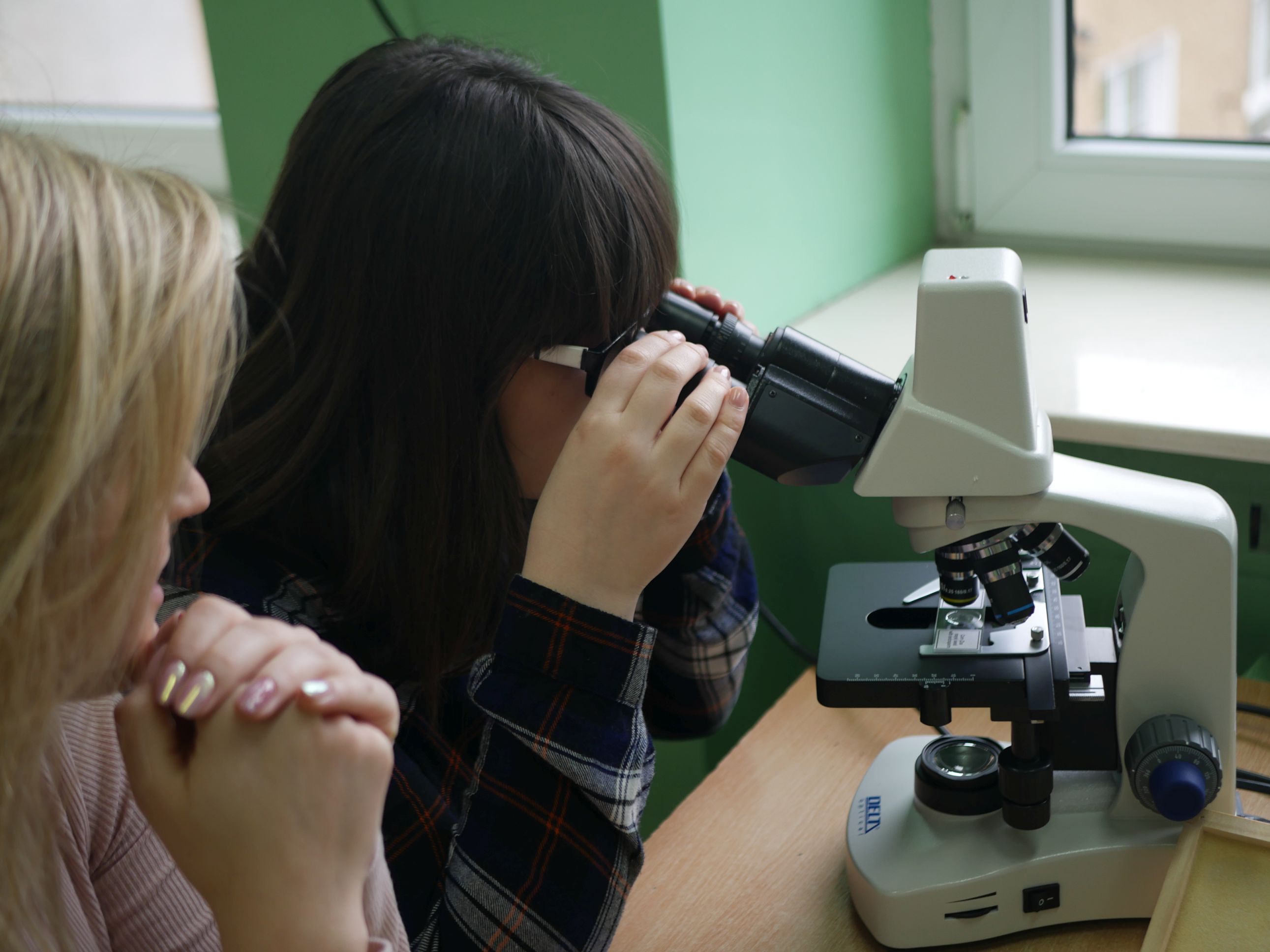 2 dziewczyny siedzące obok siebie, jedna patrzy się przez mikroskop.