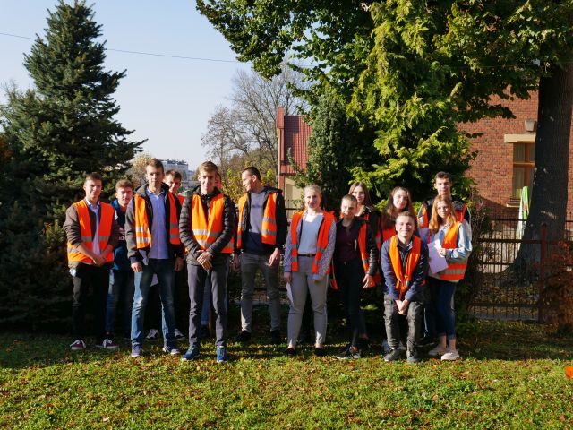 Zdjęcie grupowe uczestników olimpiady, wszyscy ubrani w pomarańczowe kamizelki odblaskowe, Zdjęcie w plenerze, w tle fragmenty domów jednorodzinnych oraz drzewa.