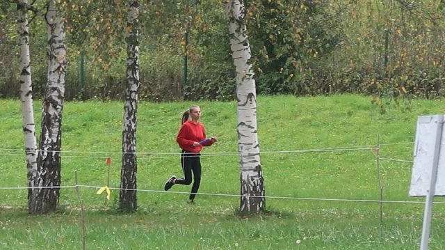 Uczestniczka w trakcie biegu. Ubrana w czerwoną bluzę, czarne spodnie oraz buty, długie blond włosy związane w koński ogon. Biegnie po soczysto-zielonej trawie, pomiędzy brzozami.