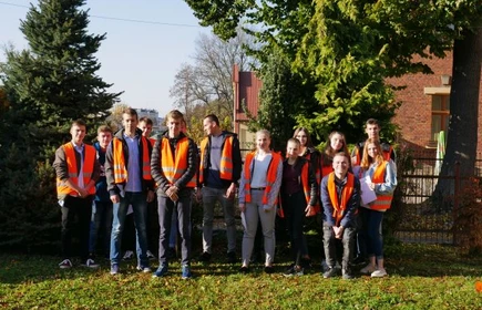 Zdjęcie grupowe uczestników olimpiady, wszyscy ubrani w pomarańczowe kamizelki odblaskowe, Zdjęcie w plenerze, w tle fragmenty domów jednorodzinnych oraz drzewa.