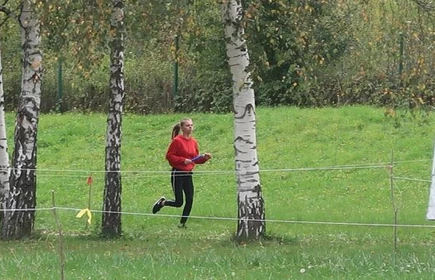 Uczestniczka w trakcie biegu. Ubrana w czerwoną bluzę, czarne spodnie oraz buty, długie blond włosy związane w koński ogon. Biegnie po soczysto-zielonej trawie, pomiędzy brzozami.