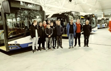 Uczniowie w hali zajezdni na tle autobusów. Obok prawdopodobnie pracownicy zajezdni. 