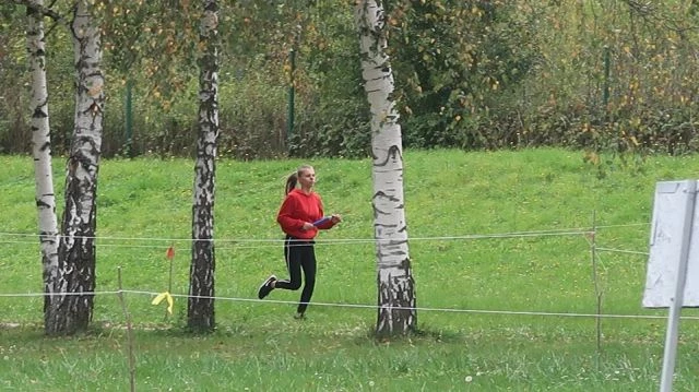 Uczestniczka w trakcie biegu. Ubrana w czerwoną bluzę, czarne spodnie oraz buty, długie blond włosy związane w koński ogon. Biegnie po soczysto-zielonej trawie, pomiędzy brzozami.